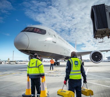 Menzies Aviation ground crew guiding an aircraft under a jet bridge during arrival on an airport apron