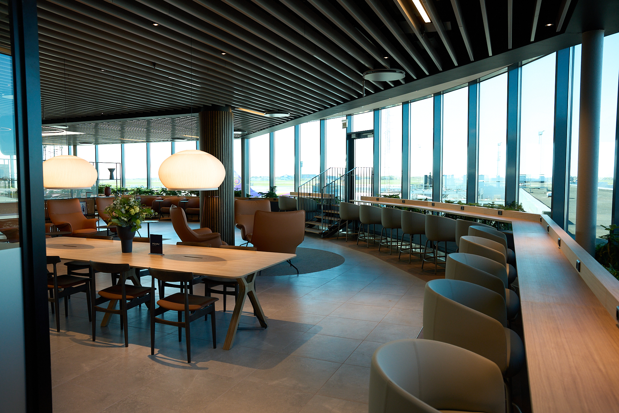 Interior of the Pearl Lounge at Copenhagen Airport with seating, communal tables and runway views through floor‑to‑ceiling windows.