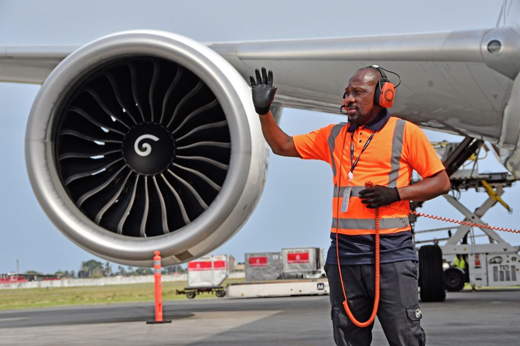 Ground crew member signalling beside a commercial aircraft engine on the airport apron.
