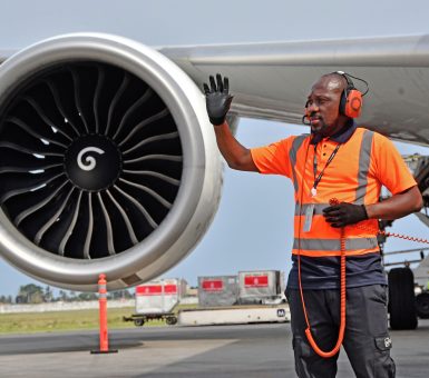 Ground crew member signalling beside a commercial aircraft engine on the airport apron.