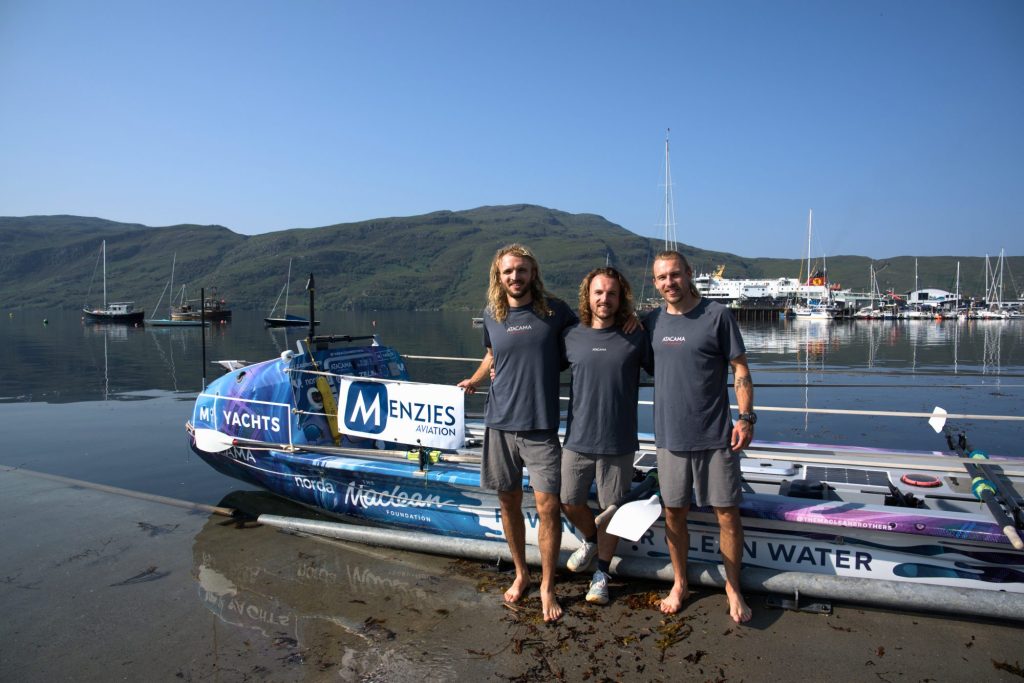 Three rowers stand beside an ocean rowing boat featuring Menzies Aviation branding at a harbour.