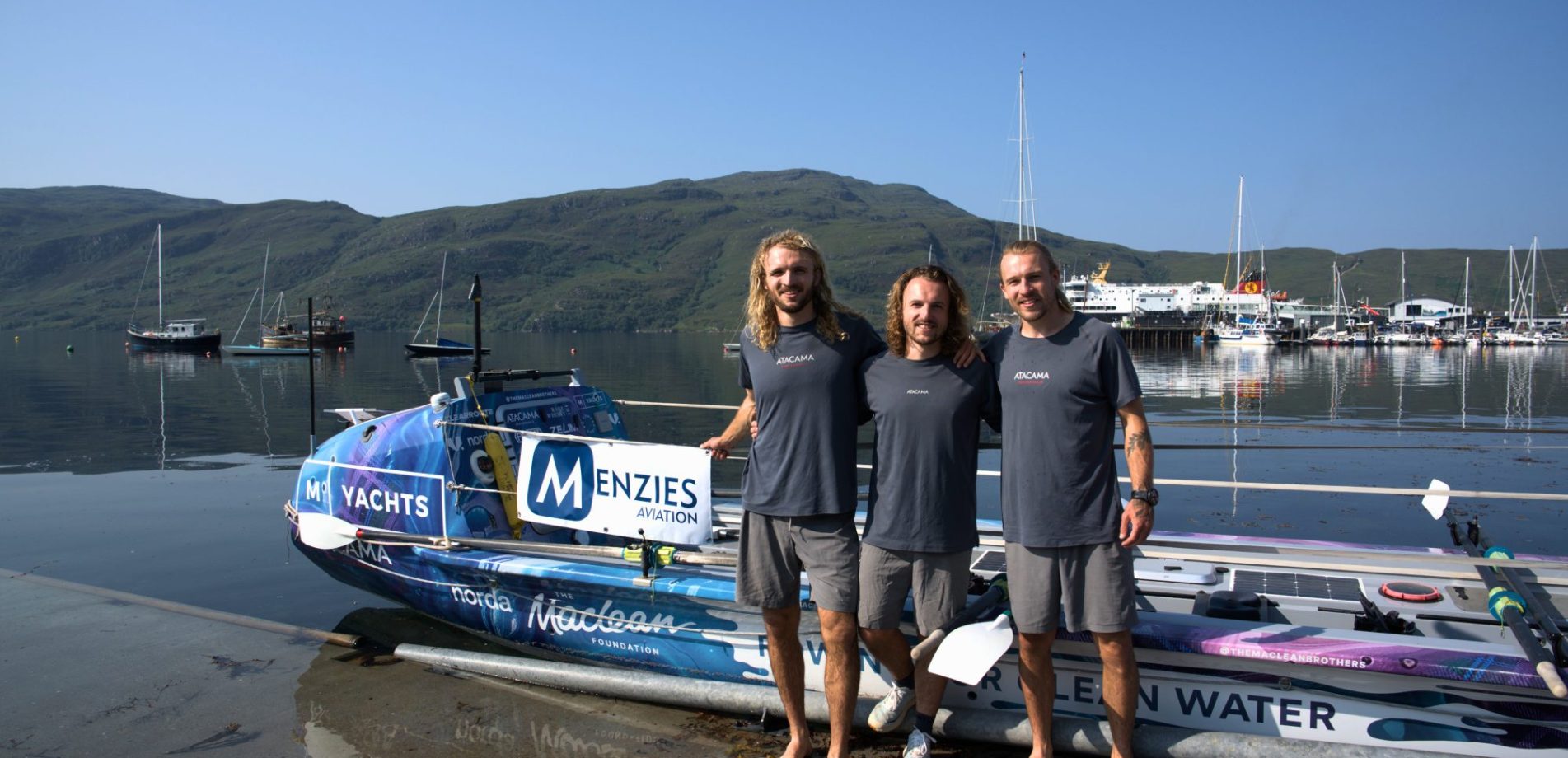 Three rowers stand beside an ocean rowing boat featuring Menzies Aviation branding at a harbour.