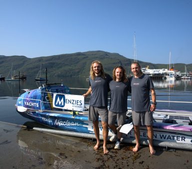 Three rowers stand beside an ocean rowing boat featuring Menzies Aviation branding at a harbour.