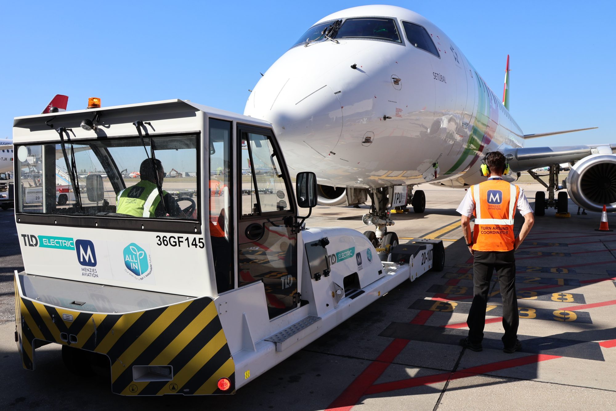 Menzies Aviation team operating an electric pushback tug alongside a TAP Air Portugal aircraft on the airport apron.